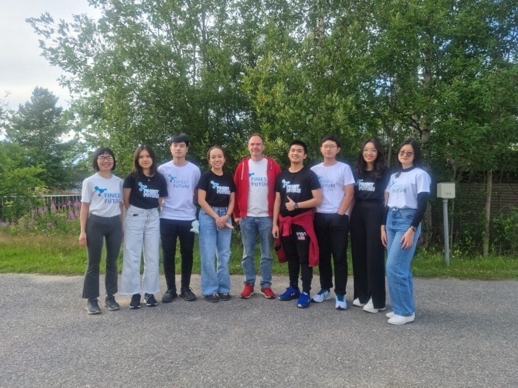 HS2021 Diary: The first Summer Camp ever in Finland! 7 A group of eight people standing side by side outdoors, smiling at the camera. They are casually dressed, mostly in jeans and T-shirts with "Finest Future" printed on them. Behind them are green trees and a cloudy sky.