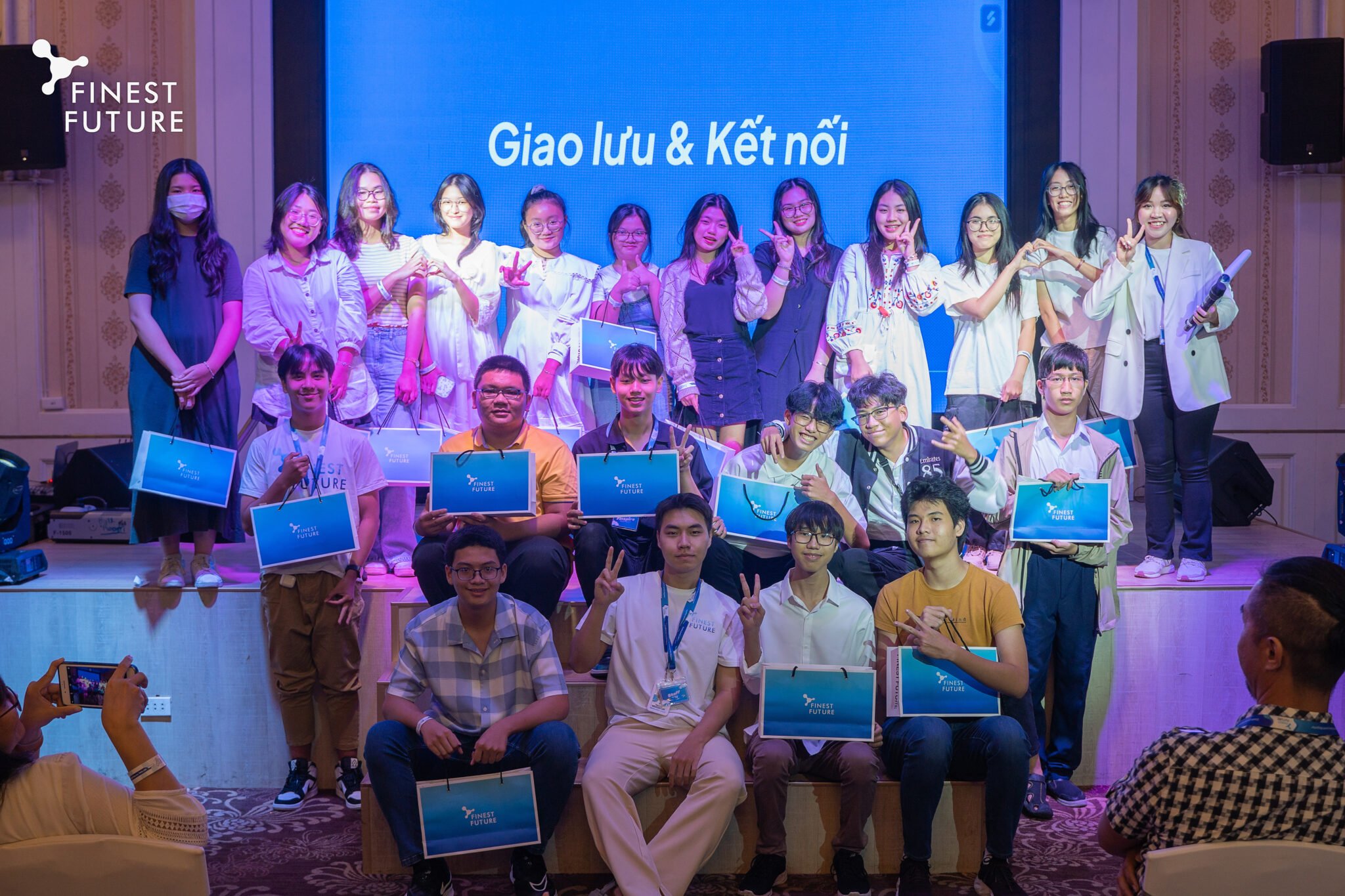 A group of young people poses on stage holding certificates and gift bags. They are smiling in front of a large screen displaying "Giao lưu & Kết nối." Some are seated, while others stand.