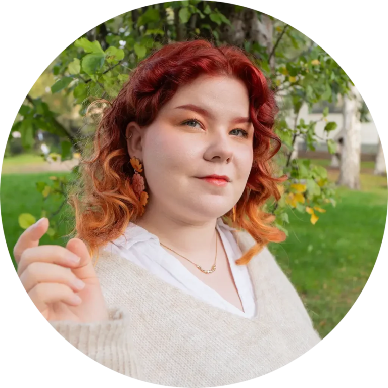 A person with curly red and orange hair stands in front of green foliage. They wear a light sweater and earrings shaped like flowers, looking toward the camera with a slight smile.
