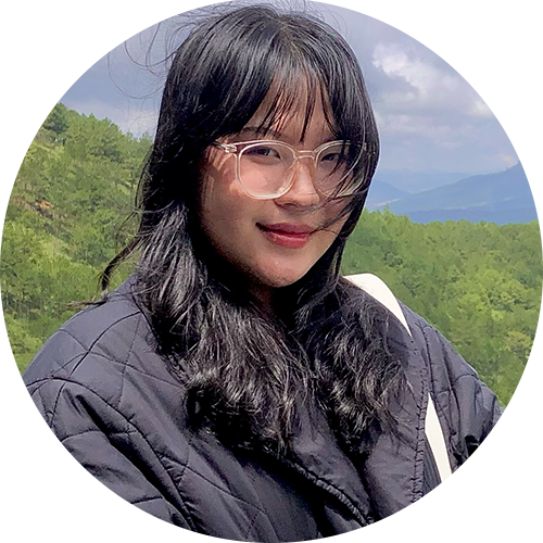 A person with long black hair and glasses, wearing a dark quilted jacket, stands outdoors. The background features lush green trees and a cloudy sky.