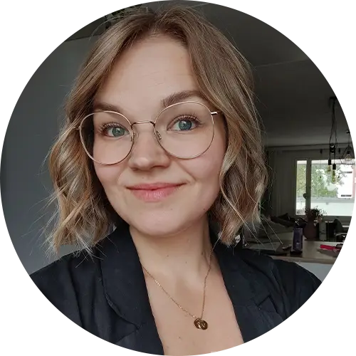 A person with shoulder-length wavy blond hair and glasses smiles at the camera. They are wearing a black top and a necklace with a round pendant. The background shows a room with a window and some plants.