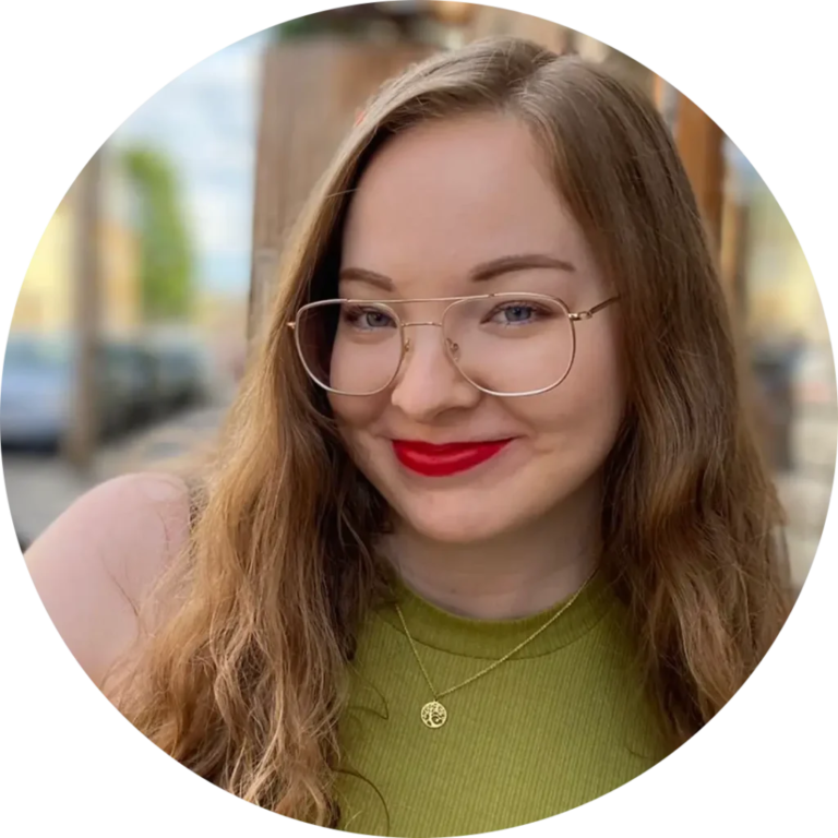 A woman with long brown hair, wearing glasses and bright red lipstick, smiles at the camera. She has on a green top and a gold necklace. The background appears to be an outdoor urban setting, slightly blurred.