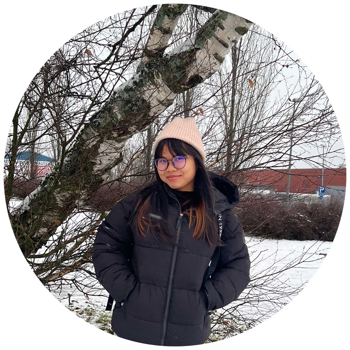 Person standing in a snowy landscape, wearing a black winter jacket and a beige beanie. They are under a tree with leafless branches, and there are buildings in the background.