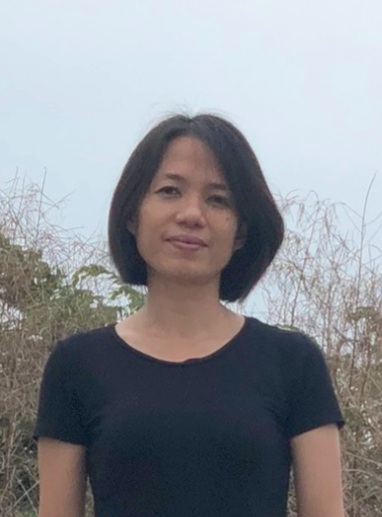 A person with short dark hair is standing outside, wearing a black t-shirt. The background includes dry plants and a cloudy sky.