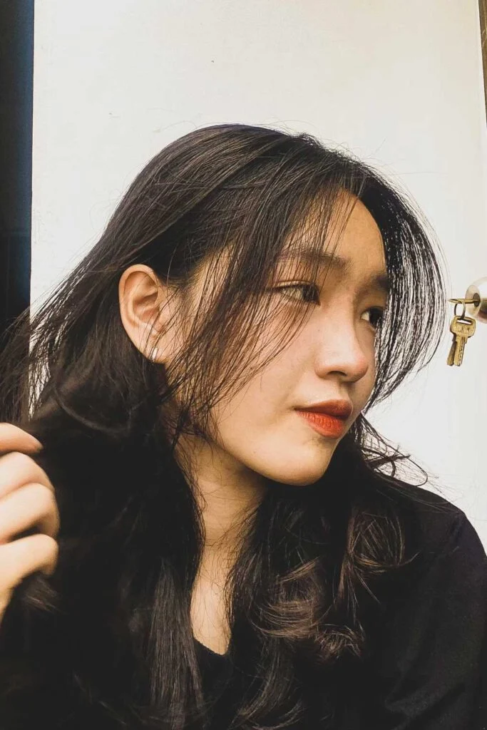A person with long, dark hair and subtle makeup looks to the side, near a white wall and a door lock. They wear a dark top, and their hand is gently holding part of their hair.