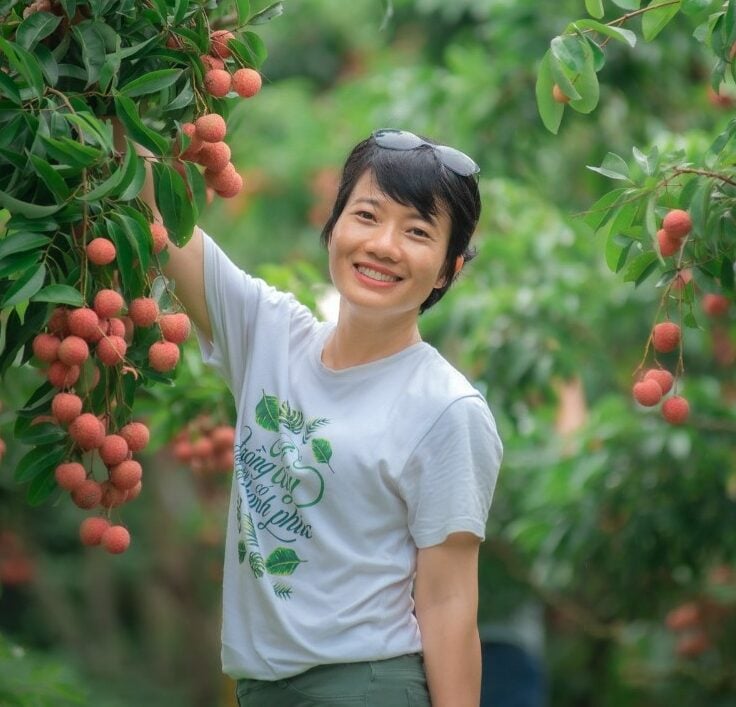 A person smiles and poses in a lush, green garden, holding a cluster of ripe lychees on the tree. They are wearing a white shirt with a printed design and have sunglasses resting on their head.