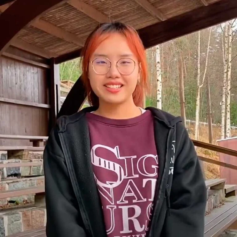 A person with orange hair and glasses smiles while standing on a wooden terrace. They're wearing a maroon shirt with white lettering and a black jacket. Trees and a wooden roof create a natural and rustic background.