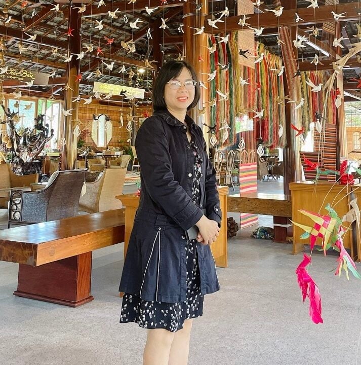 A woman in a black coat stands in a room decorated with colorful hanging origami cranes. The room has wooden furniture and vibrant fabric decorations, creating a cozy and artistic atmosphere.