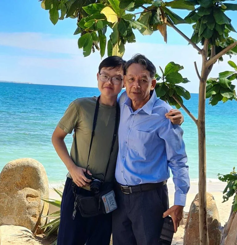 Two people stand together under a tree, smiling at the camera, with a beach and ocean in the background. One person holds a camera and the other holds a hat, enjoying a sunny day by the sea.