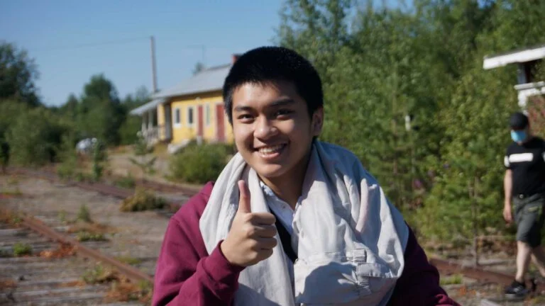 A person smiling and giving a thumbs up stands near overgrown train tracks. They are wearing a maroon sweater and a scarf. There are trees and a building in the background on a sunny day.