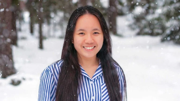 A person with long dark hair stands outdoors in the snow, wearing a blue and white striped shirt, smiling at the camera. Snowflakes are gently falling around them, and blurred trees are visible in the background.