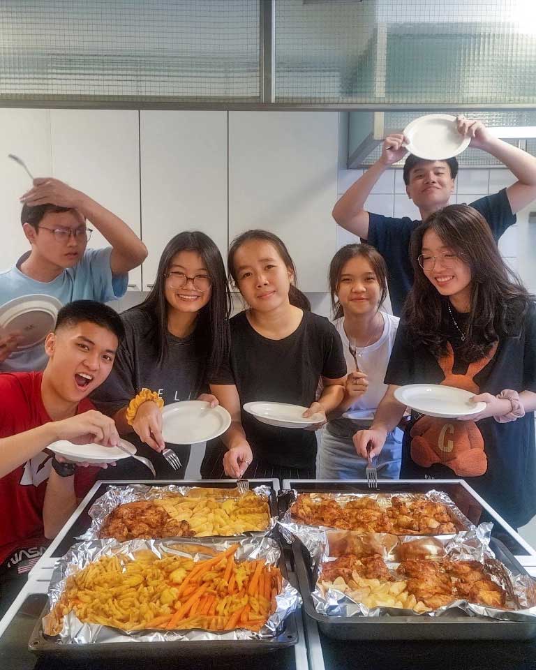 A group of seven young people gather around a kitchen counter with baked dishes of chicken, fries, and carrots. They are smiling and holding plates, ready to serve the food.
