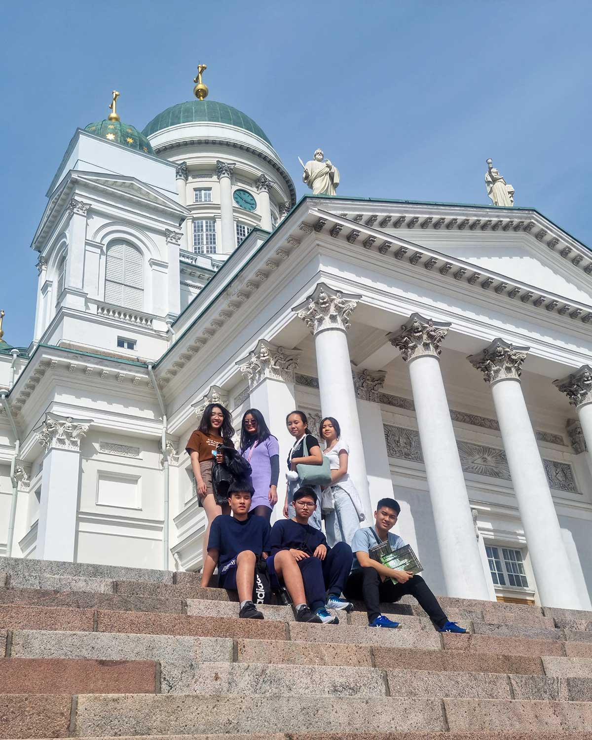 A group of seven people sitting on steps in front of a large, white neoclassical building with columns and green domes, under a clear blue sky.