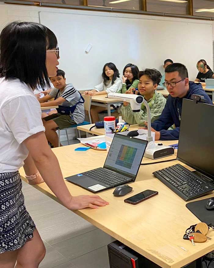 A person stands at the front of a classroom speaking to a group of students seated at desks. A laptop, smartphone, and other items are on the table. The students are listening attentively.