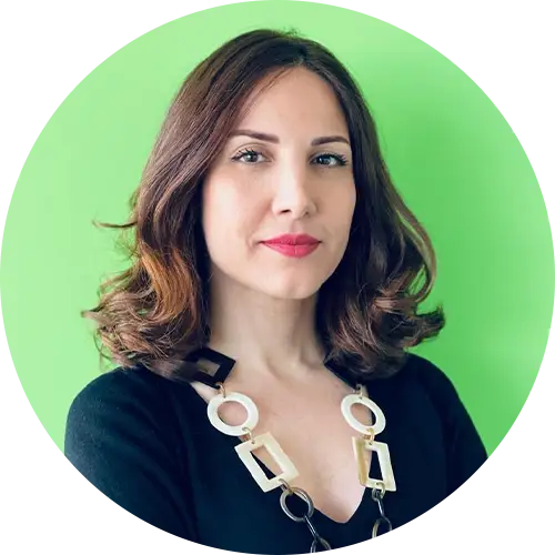A person with shoulder-length brown hair stands against a bright green background, wearing a black top and a bold, chunky necklace with rectangular links. They are looking confidently at the camera.
