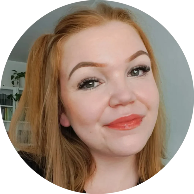 A person with long red hair smiles slightly in a circular frame. The background is soft and out of focus, with hints of greenery and books. The person has prominent eyebrows and is wearing makeup, including red lipstick.