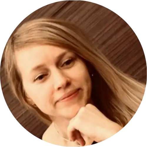 A person with long blonde hair is smiling slightly, resting their chin on their hand. The background is a wooden texture.