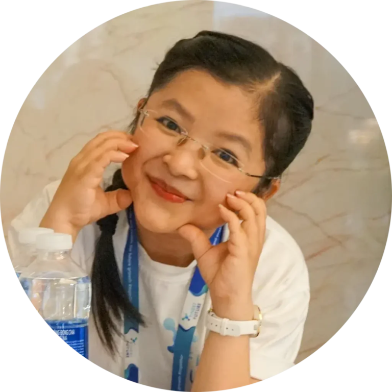 A person with braided hair and glasses smiles while playfully holding their hands near their cheeks. They are wearing a white shirt and lanyard, and a water bottle is visible in the foreground.