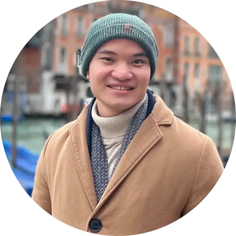 A person wearing a beige coat, gray scarf, and green beanie is smiling while standing outdoors. The background features a blurred view of buildings and boats, suggesting a waterfront or canal setting.