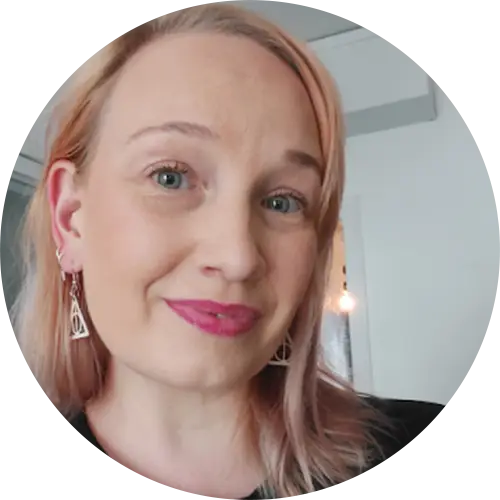 A person with shoulder-length blonde hair smiles at the camera, wearing triangular earrings and lipstick. The background is a softly lit indoor space.