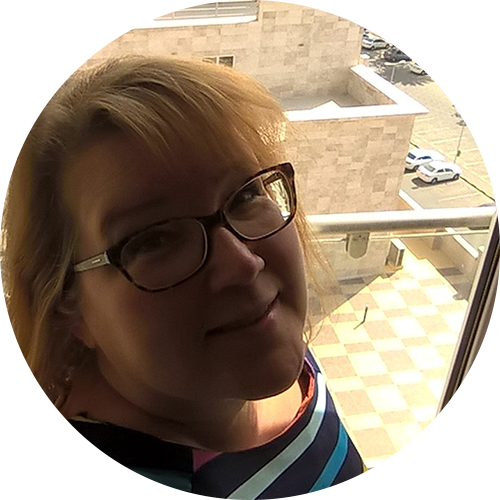 A person with glasses and light-colored hair stands by a window, smiling at the camera. The background shows a view of a tiled floor and part of a building's exterior.