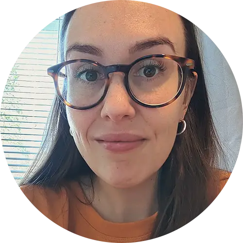 A person with long brown hair and tortoise-shell glasses is smiling at the camera. They are wearing a brown top and the background shows a window with blinds, letting in natural light.