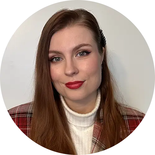 A person with long brown hair and red lipstick is wearing a red plaid jacket over a white turtleneck. The background is plain white.