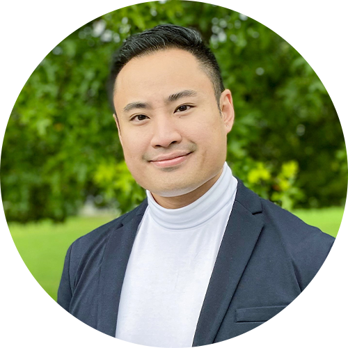 A man wearing a white turtleneck and dark blazer smiles at the camera. He stands outdoors with greenery in the background, creating a fresh and natural setting.
