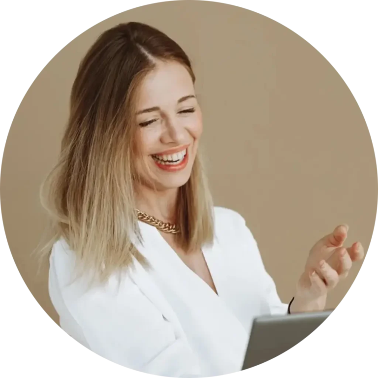 A woman with shoulder-length hair smiles while looking at a laptop screen. She is wearing a white blouse and a gold necklace, set against a plain beige background.