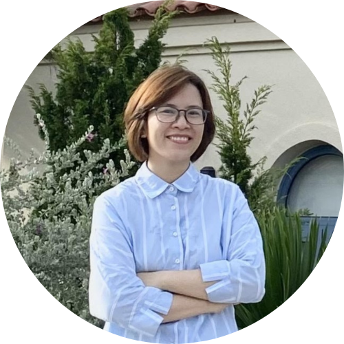 A person with short brown hair and glasses is smiling, standing outdoors in front of a building. They are wearing a light blue button-up shirt and have arms crossed. Lush green plants and a window are visible in the background.