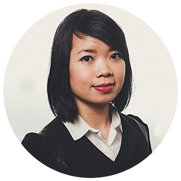 A woman with shoulder-length black hair is wearing a white collared shirt under a dark sweater. She is looking at the camera against a plain background.