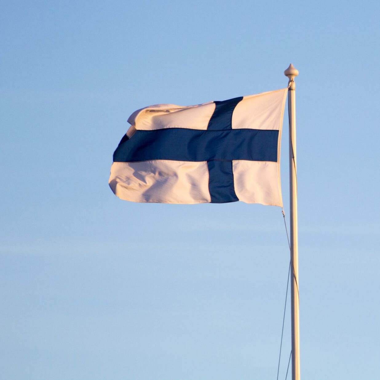 A Finnish flag with a blue Nordic cross on a white background flies against a clear blue sky.