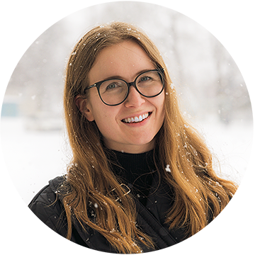 A woman with long hair and glasses is smiling while standing outdoors in a snowy setting. Flakes of snow are visible in her hair and on her dark jacket. The background is blurred with snow-covered trees.