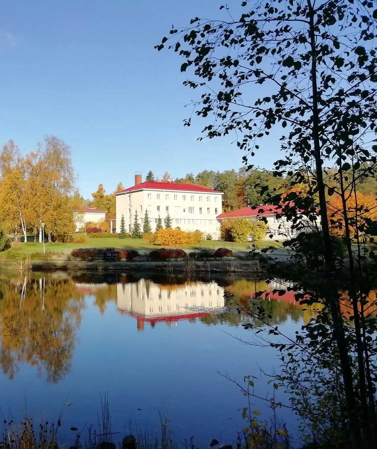 A serene autumn landscape with a white building reflecting in a calm lake. The surrounding trees have vibrant fall foliage in shades of orange and yellow, under a clear blue sky. Silhouetted branches frame the foreground.