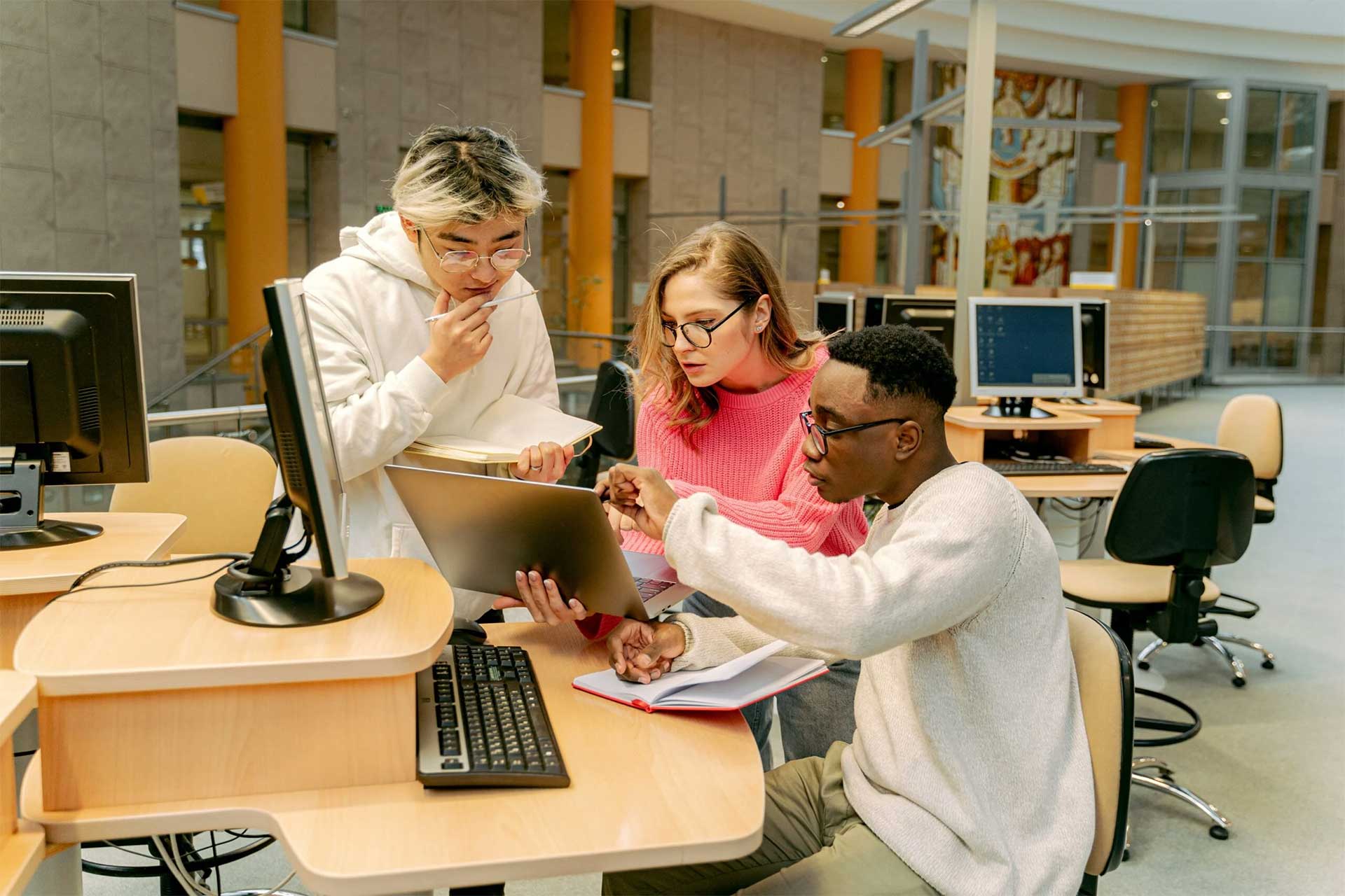 Three students collaborate in a computer lab. Two are seated while one stands, holding a laptop. They are engaged in discussion, with computers and digital equipment around them. The setting appears to be a modern educational environment.