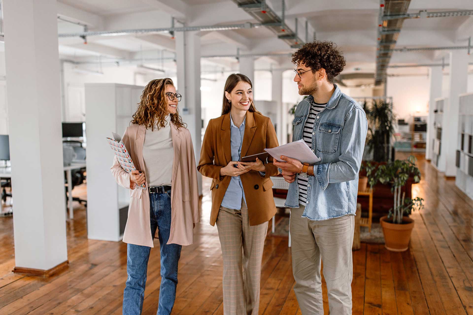 Three people walk together in a bright office space with wooden floors. They are engaged in conversation, holding documents and a tablet. The atmosphere is casual, with plants and workstations visible in the background.