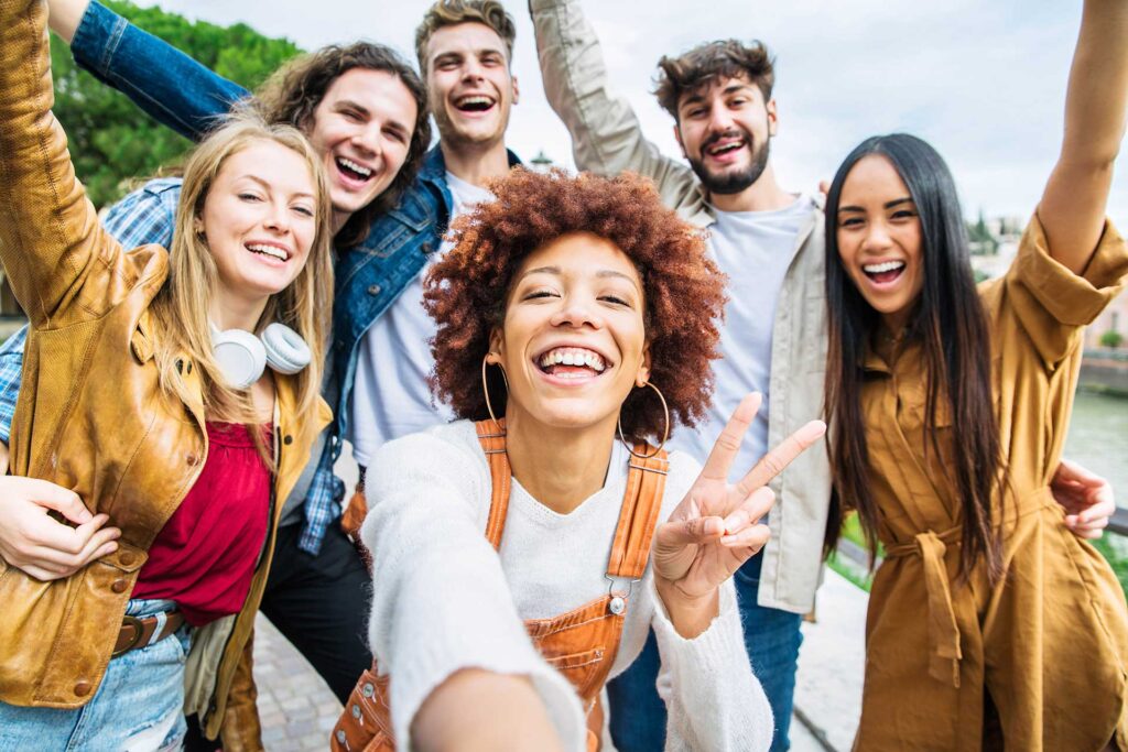 A group of six diverse young adults smiling and posing for a selfie outdoors. The person in front is making a peace sign. They are standing close together, and appear joyful and carefree, with trees and a building in the background.