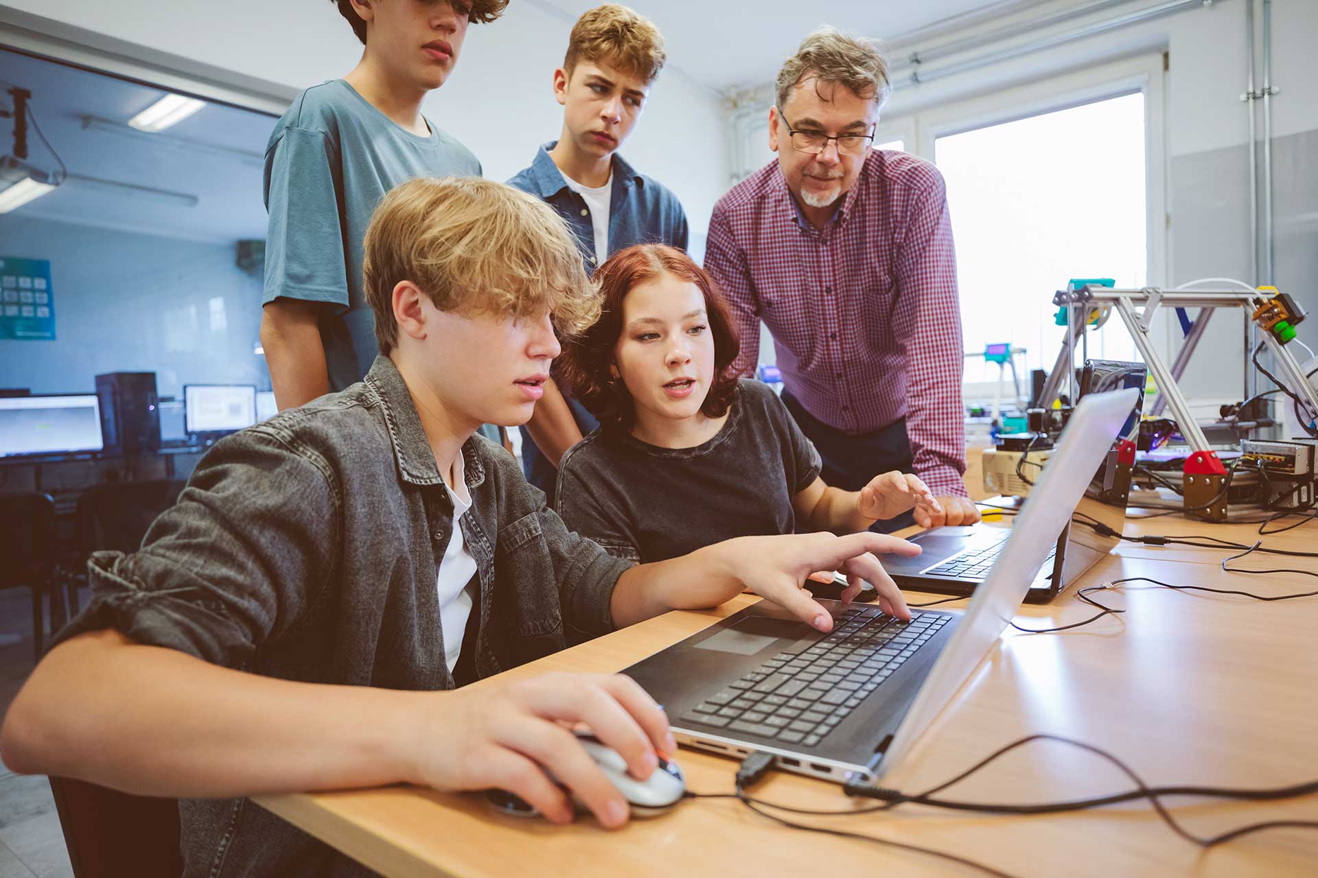 A group of students collaborates on a project using a laptop in a classroom. An adult stands nearby, guiding and observing. The setting includes a 3D printer and computers, suggesting a focus on technology or STEM education.