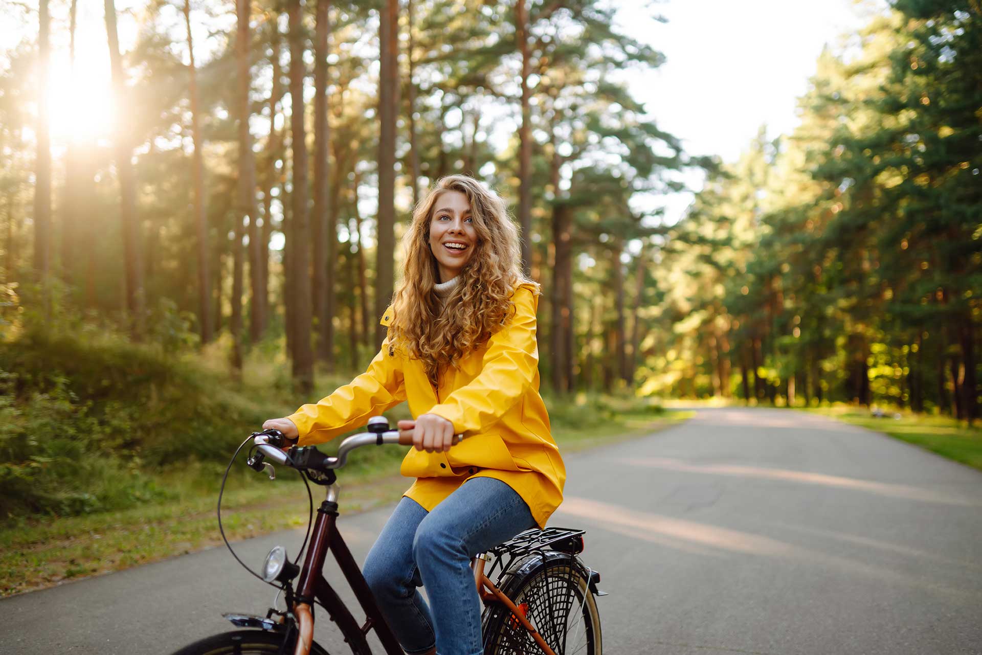 A woman in a yellow jacket rides a bicycle on a path through a sunlit forest. She is smiling and surrounded by tall trees. The atmosphere is peaceful and bright.