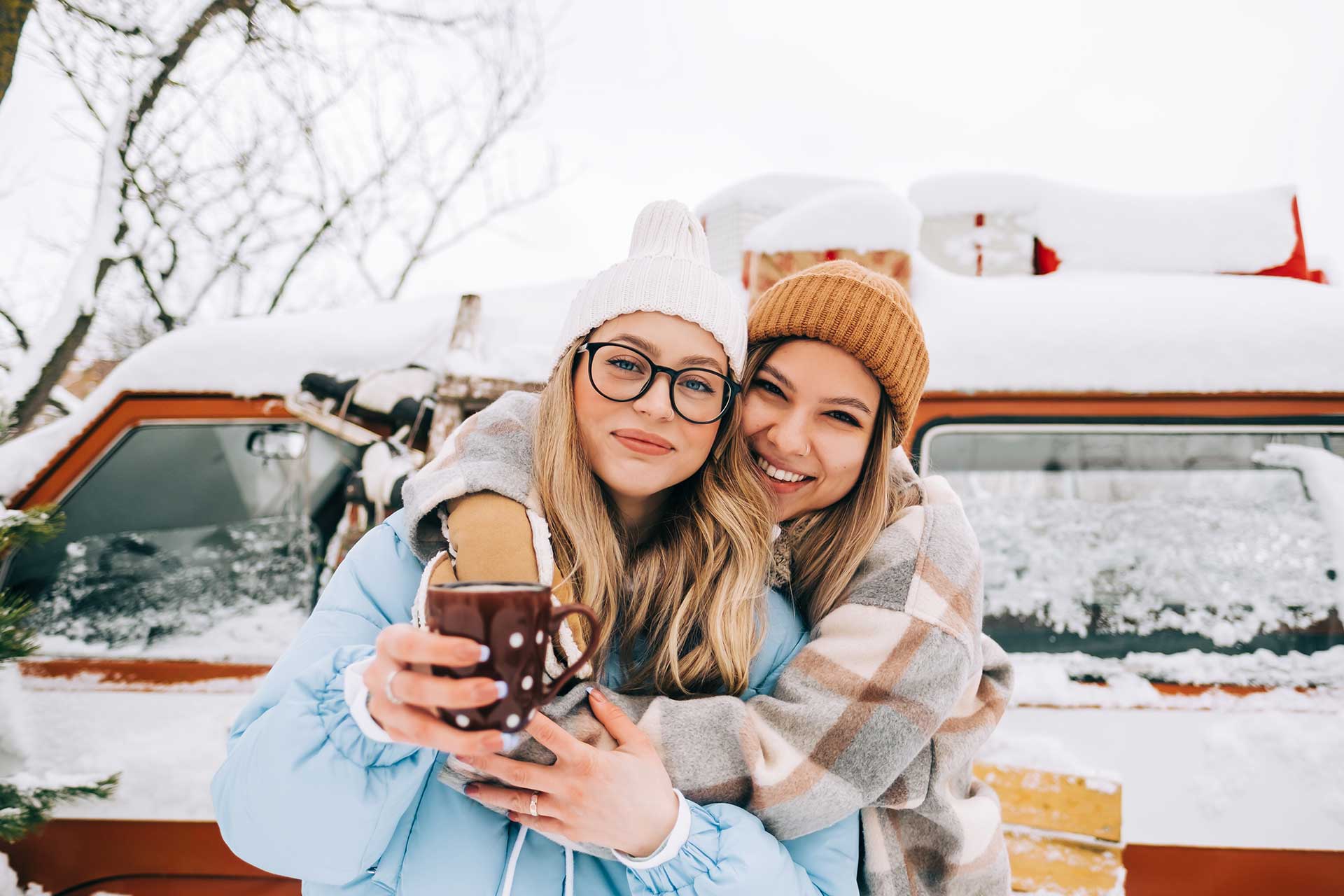 Two friends stand in front of a snow-covered van. One woman, wearing glasses and a blue jacket, holds a mug. The other, in a plaid coat, smiles and hugs her. Both wear beanies, with a snowy landscape in the background.