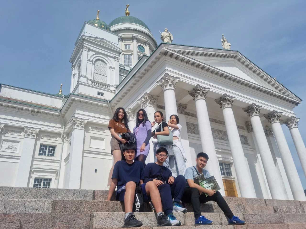 A group of six people is sitting and standing on steps in front of a large white building with columns and domes under a clear blue sky. They appear relaxed and are posing for the photo.