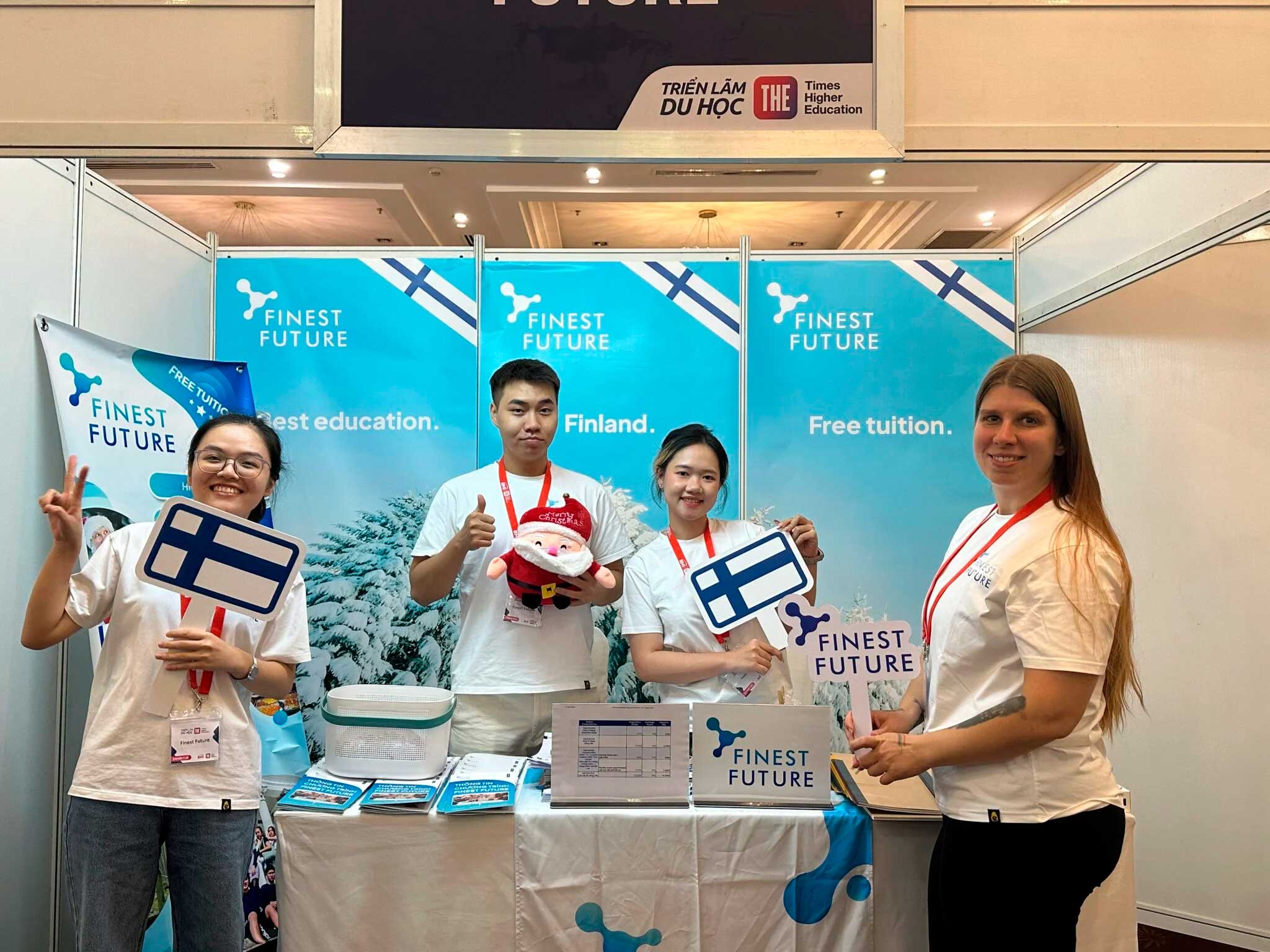 Four people smiling at a booth with "Finest Future" branding. Two hold Finnish flags, one holds a plush toy. The background features snowy landscapes and slogans like "Finest education. Finland." and "Free tuition." They wear matching white T-shirts.