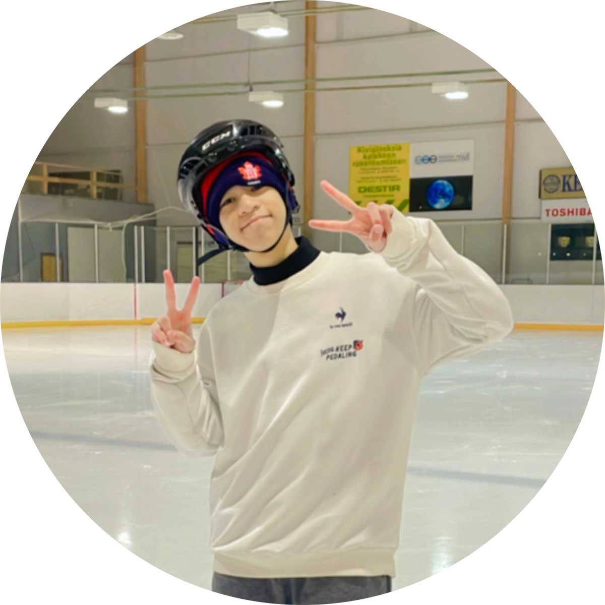 A person wearing a white sweater, black helmet, and red hat is smiling and making peace signs with both hands on an ice skating rink. The background shows the ice surface, barrier, and some advertisements.