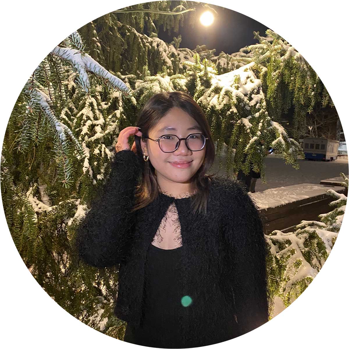 A person wearing glasses and a black outfit stands smiling in front of snow-covered evergreen branches at night, illuminated by a nearby streetlight.
