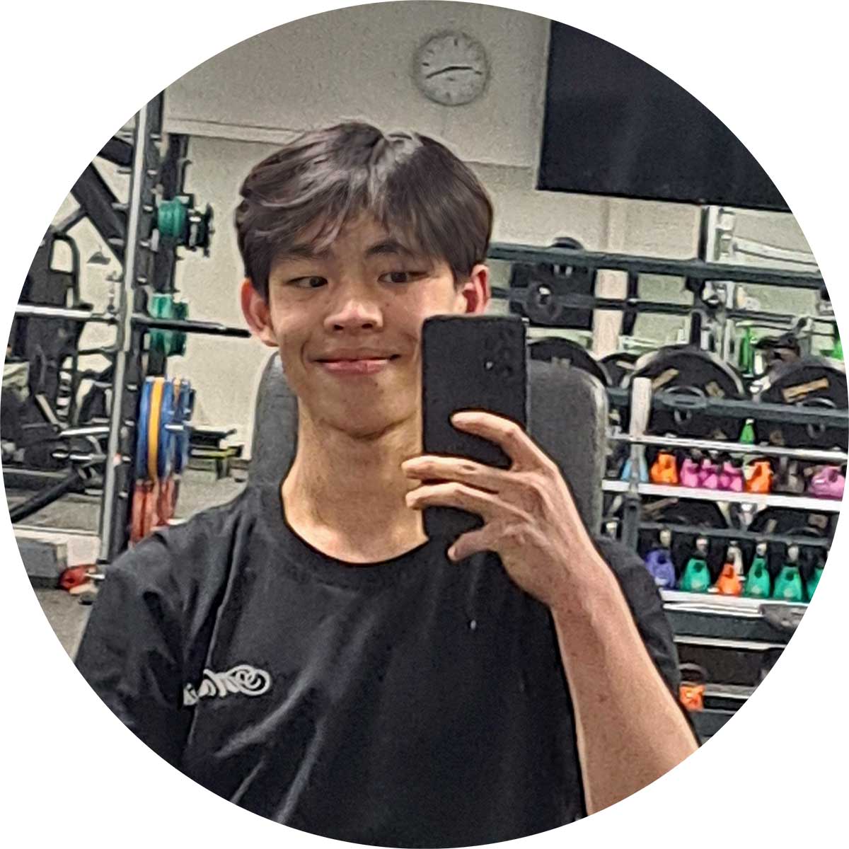 A person taking a selfie in a gym with various weights on racks in the background. They are holding a smartphone, wearing a black shirt, and standing in front of a mirror.