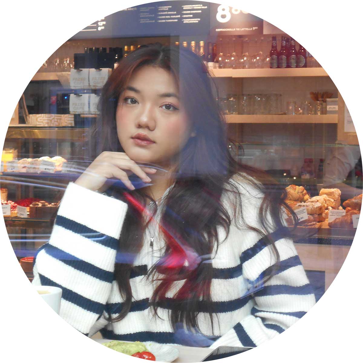 A person with long, dark hair sits inside a cafe, wearing a striped sweater. They rest their chin on their hand, looking out the window, with reflections of the cafe's interior visible on the glass.