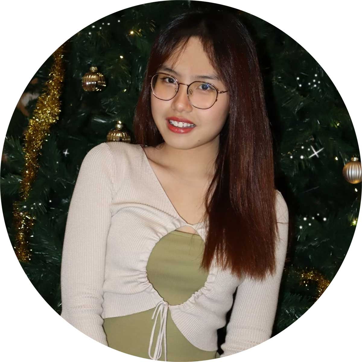 A person with long brown hair and glasses smiles in front of a Christmas tree adorned with gold tinsel and ornaments. They are wearing a light-colored top with a ruched front.