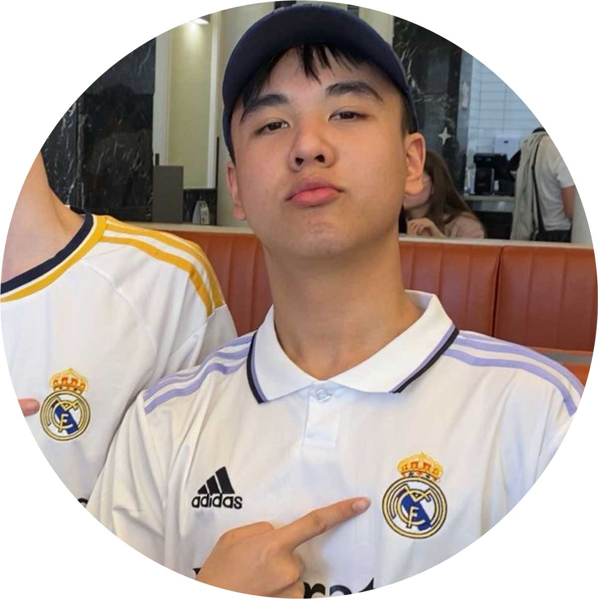 A person wearing a Real Madrid soccer jersey and a cap points to the team logo on their chest. They are seated indoors with a blurred background.