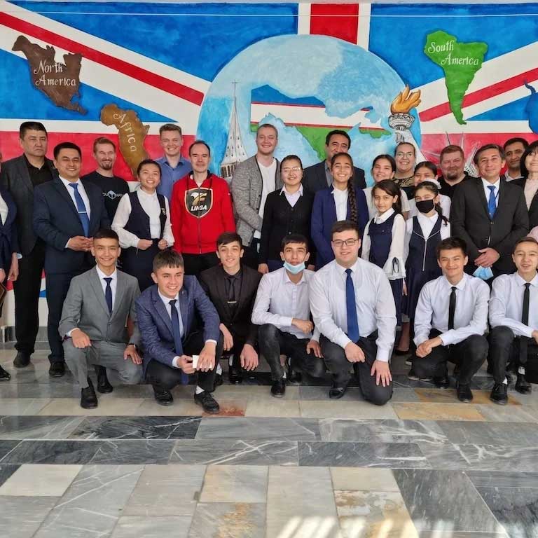 A diverse group of people, dressed in formal and casual attire, pose in front of a colorful mural depicting world maps and flags. Some are standing while others kneel in front, smiling at the camera.