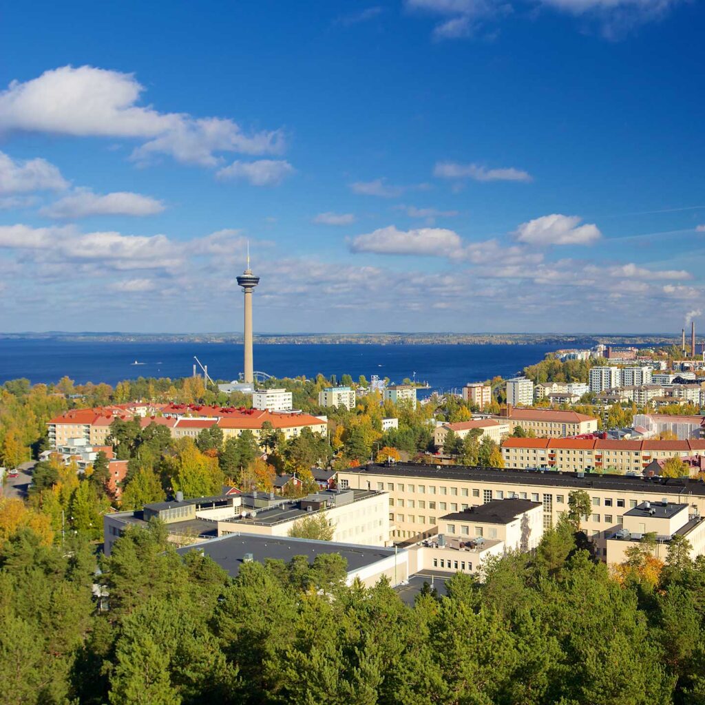 A scenic view of a city with a tall observation tower near a large lake. The landscape includes a mix of buildings, trees with fall foliage, and a clear blue sky with scattered clouds.